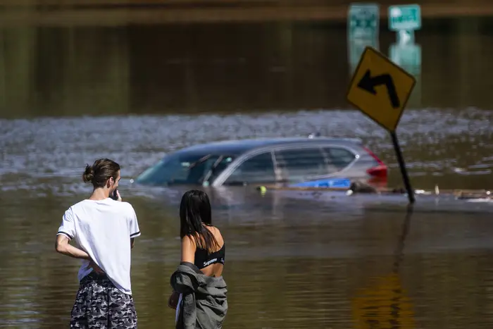 An SUV is submerged in flood water in the distance as two people look at it